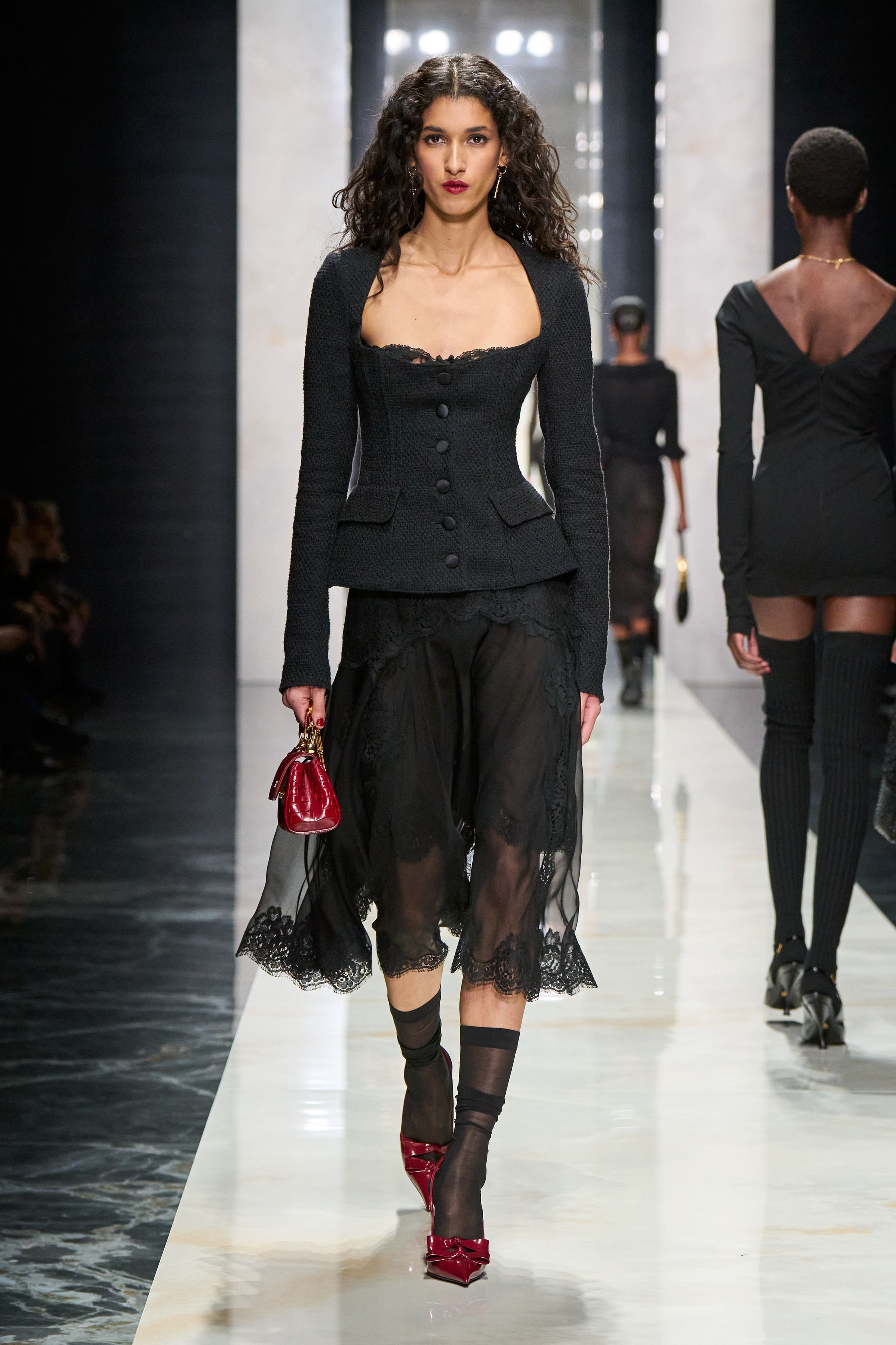 Dolce & Gabbana在本季回归品牌标志性的性感意大利美学，通过黑色蕾丝、修身西装与透视礼服塑造极具戏剧张力的女性形象。系列以黑色为核心色调，结合紧身胸衣结构、透明蕾丝裙与宽肩西装，形成刚柔并济的视觉语言。皮草外套与复古帽饰则增添奢华氛围，使整体造型在性感与优雅之间保持平衡，呈现出典型的意大利电影般的华丽气质-顽石频道趋势研究